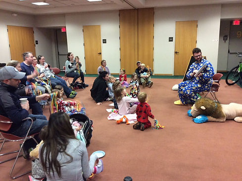 A librarian in playful blue pajamas holds a book and reads to a group of children and adults surrounding him at storytime