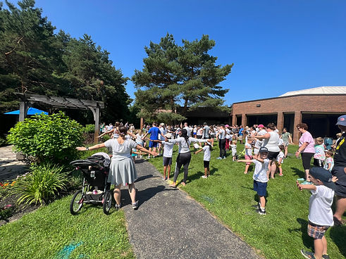 A large group of parents and children all hold their hands out in a yoga pose in the orion library reading garden located at the back outdoor area of the library. The library is visible behind the group of patrons.