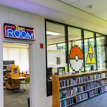 the teen room at orion library with pokemon art on the windows, a shelve of books before the entrance, and a neon Teen Room sign above the rooms entrance