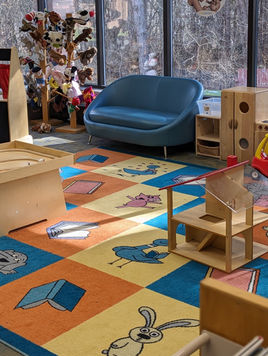 The youth room at Orion Library with a chair next to a window where parents can sit and a rug with playful cartoons, and toys for kids to play with. 
