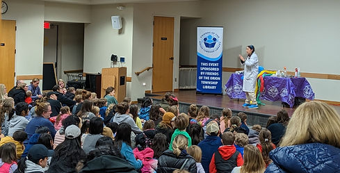 A presenter in a science lab coat in the meeting room at the library stands on the stage presenting science experiments as a group of parents and children sit on the floor and watch